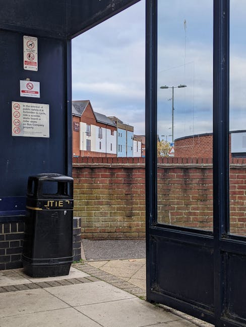 Inside a shelter with dark blue metal walls, a black plastic rubbish bin with a slightly domed lid is positioned in the left corner. The bin features yellow text or signage partially visible on its su