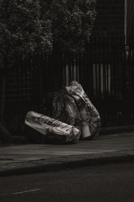 Two large garbage bags filled with waste material are positioned on the pavement near a black metal fence with vertical bars. The bags appear to be made of thick plastic and are loosely tied at the to
