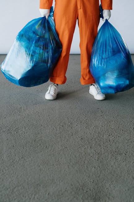 Two large garbage bags filled with waste material are positioned on the pavement near a black metal fence with vertical bars. The bags appear to be made of thick plastic and are loosely tied at the to