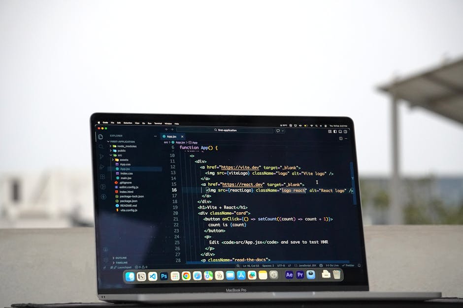 A laptop computer with a silver metallic finish and a black screen displaying a code editor containing lines of JavaScript and HTML code in various colors, mounted on a light-colored surface against a